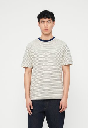 Ung mand med sort hår iført hvid T-shirt med korte ærmer og tynde sorte striber samt en mørkeblå krave, kombineret med mørkeblå jeans.