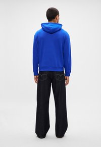 Férfi kék kapucnis pulcsiban és széles szárú fekete farmert viselve, háttal áll egy sima fehér háttér előtt.