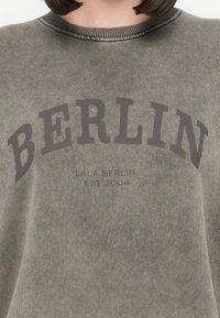 Felpa verde oliva con collo a costine, caratterizzata dalla scritta "BERLIN" in grigio scuro in un testo arcuato e "LALA BERLIN EST 2004" sottostante. Tessuto morbido.