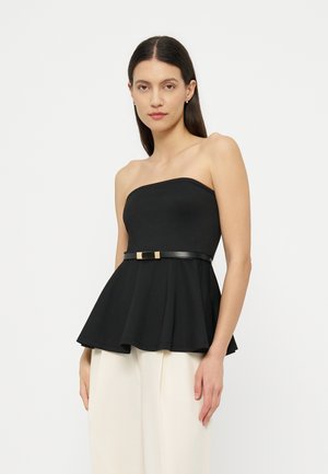 Γυναίκα που φορά ένα μαύρο τοπ πεπλούμ χωρίς τιράντες με λεπτή ζώνη και λευκό παντελόνι με φαρδιές παντελόνες, στέκεται μπροστά σε απλό φόντο.