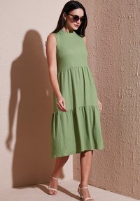 Light green sleeveless dress with a textured fabric, tiered design, and knee-length silhouette; paired with open-toe sandals.