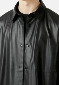 Chaqueta de camisa de cuero negro con una textura suave, cierre de botones, cuello puntiagudo y un bolsillo frontal. Presenta botones de presión metálicos.