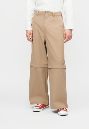 Pantalon convertible beige avec jambes supérieures amovibles zippées, porté avec des baskets basses rouges et une chemise blanche à manches longues sur une personne debout.