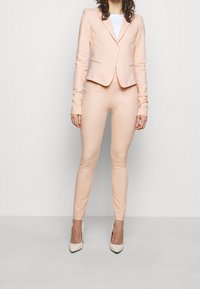 Koralowy żakiet na miarę z długimi rękawami i zapięciem na jeden guzik, zestawiony z dopasowanymi, wąskimi spodniami. Gładka tekstura, minimalistyczne detale.