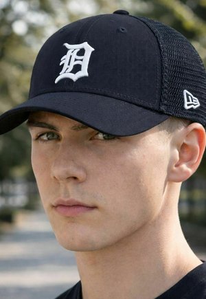 Junger Mann, der eine schwarze Baseballkappe mit weißem Old English "D"-Logo und Netzrücken trägt, im Freien vor unscharfem Baumhintergrund.