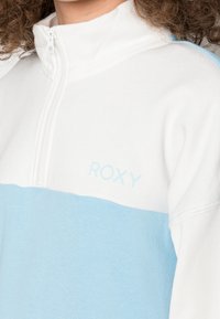Halbzip-Pullover aus weißem und hellblauem Fleece. Mit einem Roxy-Logo in Hellblau, geripptem Kragen und weichem Textil.
