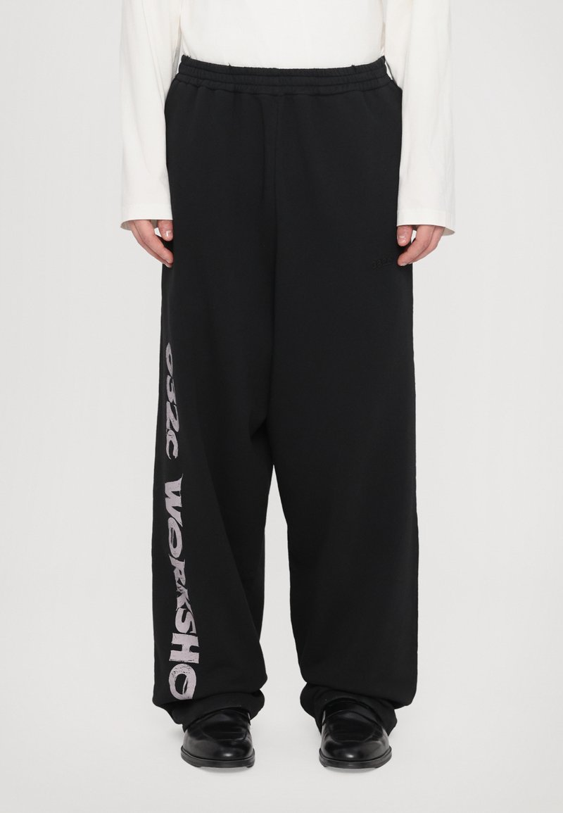 Schwarze Sweatpants mit entspannter Passform, elastischem Bund und dem Text "9926 WORKSHOP" in Weiß auf dem linken Bein. Glatte Textur.