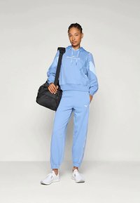 Νεαρή γυναίκα σε ανοιχτό μπλε φόρμα και λευκά αθλητικά παπούτσια στέκεται κρατώντας μια μαύρη τσάντα γυμναστηρίου στον έναν ώμο μπροστά σε ένα απλό φόντο.