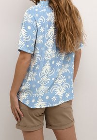 Blå kortärmad skjorta med ett vitt blommönster och rynkad halsringning, ihop med ljusbruna uppvikta shorts.