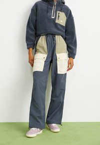 Fleece-hoodie i mörkblått med halvdragkedja och framficka; kombinerad med grå cargobyxor med krämfärgade lappar och guldtrimm.
