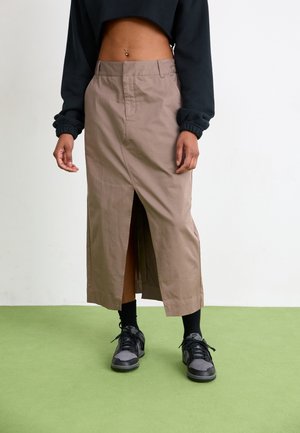 Personne portant une jupe midi beige avec une fente à l’avant, un sweat-shirt court noir, des chaussettes noires et des baskets noires et grises, debout sur un sol vert.
