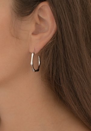 Boucle d'oreille en argent en forme irrégulière et torsadée, avec une texture polie, suspendue à un petit support sur l'oreille d'un modèle.