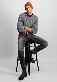 Grå langermet V-hals genser, mørke jeans, brun lærbelte, brune Chelsea-boots. Modell sitter på en svart krakk med en strikket brun beanie.