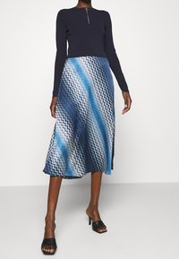 Kvinna i en marinblå långärmad topp med en framdragbar dragkedja och en blå zigzag-mönstrad midiklänning, kombinerad med svarta klacksandaler.