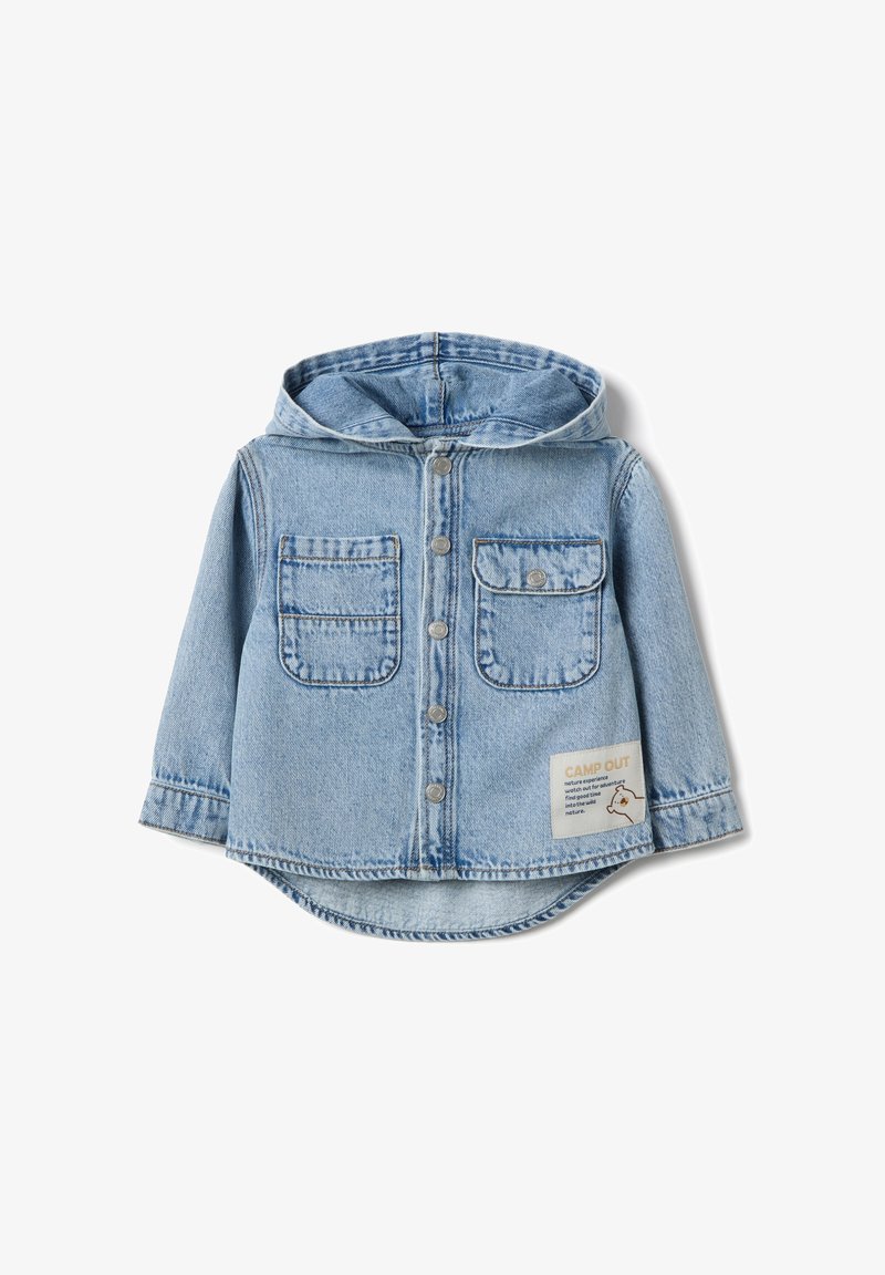 Camisa con capucha de mezclilla azul claro con cierre de botones, dos bolsillos en el pecho y una etiqueta de tela con "CAMP OUT" en el costado inferior. Textura suave.