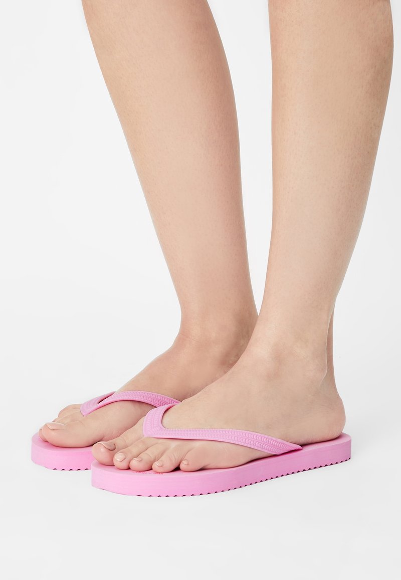 Pieds portant des tongs roses avec des brides texturées et des semelles épaisses, présentés sur un fond blanc uni.