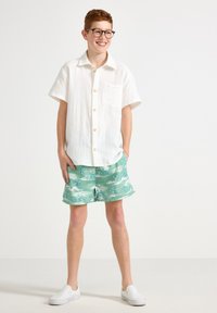 Camicia bianca di lino a maniche corte con chiusura a bottoni, abbinata a pantaloni da bagno verdi chiari con motivo e scarpe slip-on bianche.