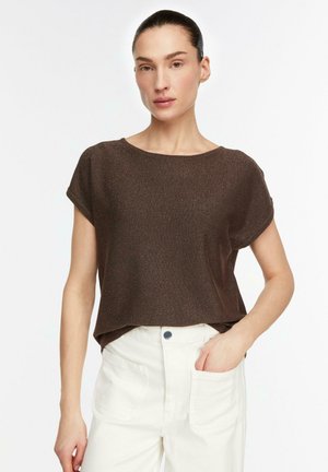 Frau mit dunkelbraunem kurzärmeligem Oberteil, das locker in hochtaillierte weiße Hose gesteckt ist, stehend mit einer Hand in der Tasche vor weißem Hintergrund.