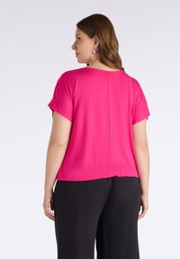 Top magenta de manga corta con escote redondo y corte holgado, visto desde la espalda, combinado con pantalones negros. Textura de tela suave.