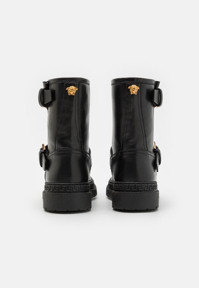 Bottines en cuir noir avec des accents de tête de lion en or, des détails de sangle et une semelle en caoutchouc noir texturé avec un motif géométrique.