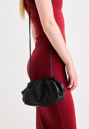 Black, slouchy handbag made of smooth material, featuring a drawstring closure and two shoulder straps, held by a person wearing a red dress.