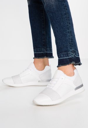 Hvide lave sneakers med elastisk bånd og snøredetalje, båret med mørke jeans med rå kanter på hvid baggrund.