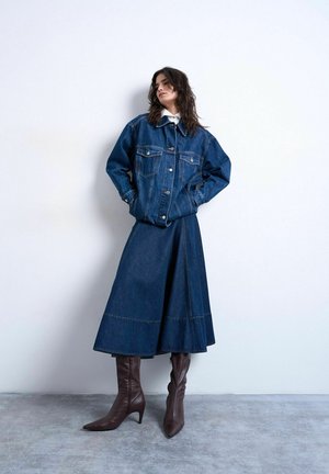 Vrouw draagt een donker denim jack en bijpassende midirok met bruine laarzen met hak, staand met handen in de jaszakken tegen een witte muur.