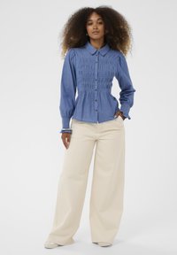 Vaaleansininen farkkupaita, jossa on kerätty etuosa, napitus, puffihihat ja elastiset hihansuut, yhdistettynä vaalean kermanvärisiin leveälahkeisiin housuihin.