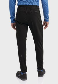 Schwarze leichte Hose mit elastischem Bund, schmaler Passform und Bündchen. Auffälliges Logo-Detail am unteren Bein. Getragen mit schwarzen Schuhen.