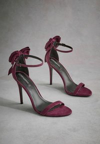 Talons hauts en suédine bordeaux avec un bout ouvert, une fine lanière à l'avant et un accent en nœud à la cheville. Talon aiguille mince.