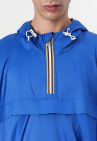 Blå fleecejacka med huva, med en kort dragkedja och flerfärgade ränder längs dragkedjan. Lättviktigt material och avslappnad passform.