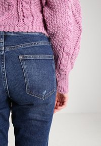 Růžový, texturovaný svetr s kabelovým pletením kombinovaný s tmavě modrými džínami obsahujícími zadní kapsy a mírné opláchnutí blízko švu.
