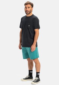 Black cotton t-shirt with a chest pocket, paired with teal shorts and black sneakers with white soles and black socks featuring a logo.