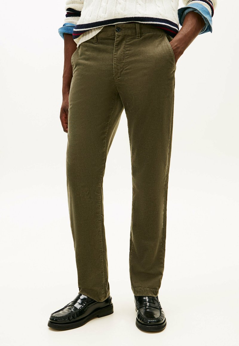 Uomo che indossa pantaloni in velluto a coste verde oliva, mocassini neri lucidi e un maglione color crema con bordi blu navy e rossi, mani in tasca in modo disinvolto.
