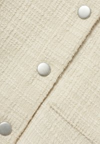 Tela color crema con un patrón texturizado, que presenta botones metálicos plateados. La trama es suave con líneas verticales y detalles en relieve.