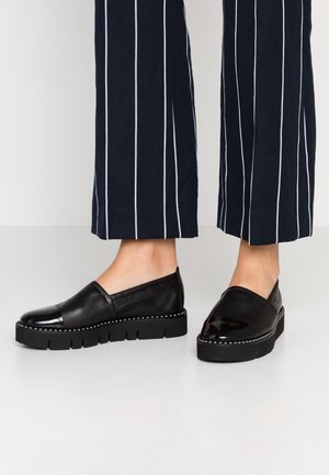 Sorte læder slip-on sko med klodsede såler og nittebesætning, båret med navyblå bukser med hvide lodrette striber.