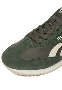 Zielony but sportowy wykonany z zamszu i nylonu, z zaokrąglonym noskiem, fakturowaną powierzchnią, sznurowaniem i kontrastową beżową podeszwą.