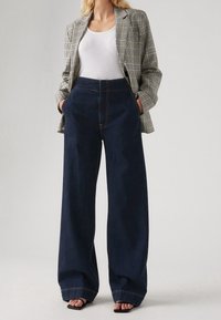 Mujer vistiendo un top blanco sin mangas, un blazer de cuadros, jeans de pierna ancha de tiro alto en azul oscuro, y sandalias negras de dedo al aire, con las manos en los bolsillos.