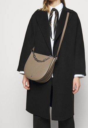 Tmavě hnědá kožená ramenní taška s zakřiveným tvarem, stříbrným řetízkovým zdobením a hladkou texturou, nošená s černým kabátem a bílou košilí.