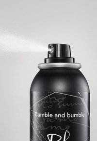 Svart cylindrisk sprayburk med en silverfärgad munstycke, med en texturerad etikett som har vit text och abstrakta mönster. Spraydimma är synlig.