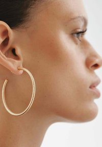 Pendiente de aro en tono dorado con textura acanalada, de gran forma circular y acabado suave, llevado en una oreja sobre un fondo claro.