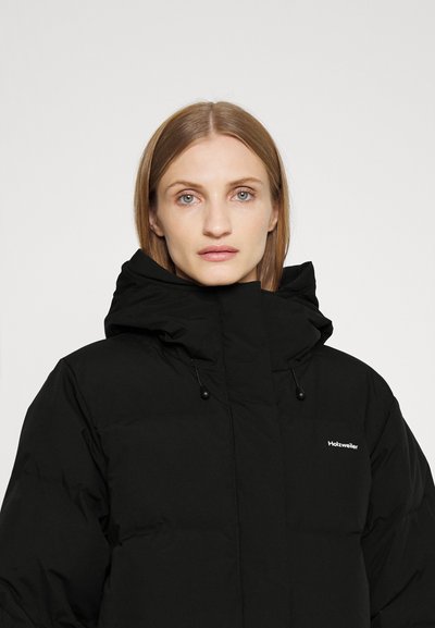 Crna pufast jakna s visokim ovratnikom, s kapuljačom sa konopcima i suptilnim logotipom brenda na prsima. Glatka nylon tekstura, oversized kroj.