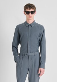 Camisa gris de manga larga con cuello abotonado, combinada con pantalones gris ajustados que presentan un cinturón y gafas de sol rectangulares negras.