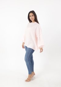 Jasnoróżowy, oversize sweter z wysokim dekoltem i szerokimi rękawami, w zestawieniu z dopasowanymi niebieskimi dżinsami oraz przezroczystymi sandałami na słupku.