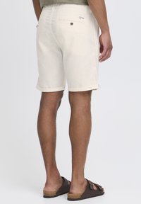 Shorts de algodón color beige con dobladillos, dos bolsillos traseros y un logo en la cinturilla. Llevados con sandalias marrones sobre un fondo neutro.