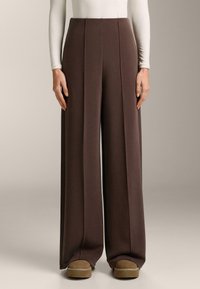 Hlače širokih nog v bogatem rjavo rjavem tkanju, z navpičnimi šivi in gladko teksturo, kombinirane s svetlimi čevlji.
