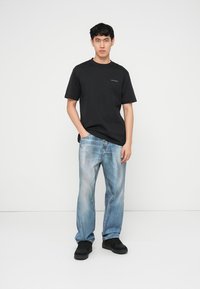 Νέος άνδρας που στέκεται, φορώντας μαύρο T-shirt, ξεβαμμένο μπλε τζιν και μαύρα αθλητικά παπούτσια, μπροστά σε απλό λευκό φόντο.