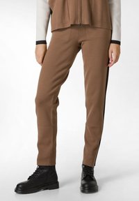 Bruine broek gemaakt van een zachte, gestructureerde stof met een getailleerd ontwerp, met zijdelen en een contrasterende zwarte streep. Gestyled met zwarte laarzen.