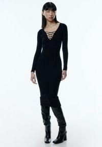 Schwarzes, geripptes Langarmkleid mit tiefem V-Ausschnitt und Schnürdetail. Kombiniert mit kniehohen, schwarzen Lederstiefeln mit quadratischer Zehenform.