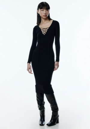 Schwarzes, geripptes Langarmkleid mit tiefem V-Ausschnitt und Schnürdetail. Kombiniert mit kniehohen, schwarzen Lederstiefeln mit quadratischer Zehenform.